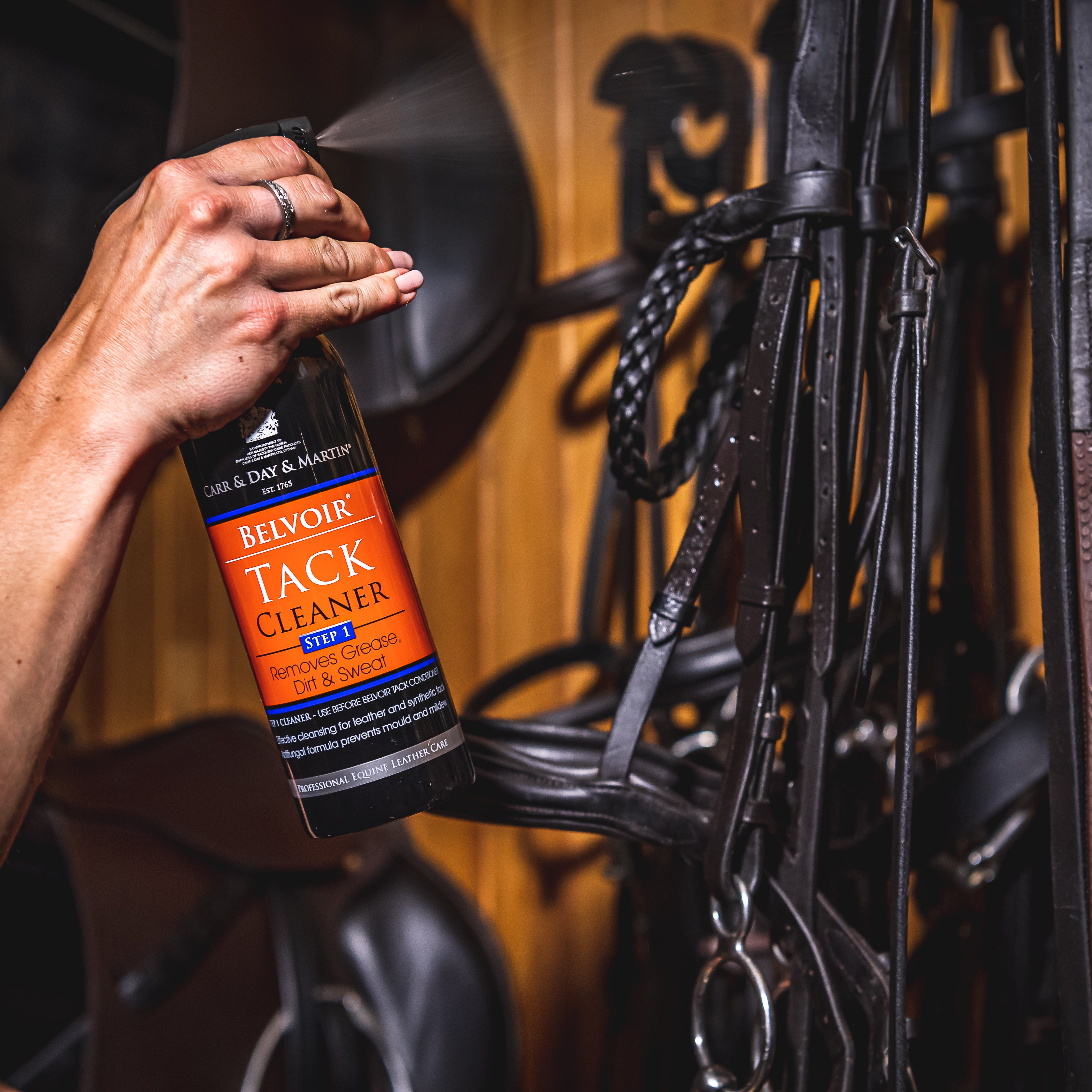 Hand holding a bottle of Tack Cleaner in front of horse tack.
