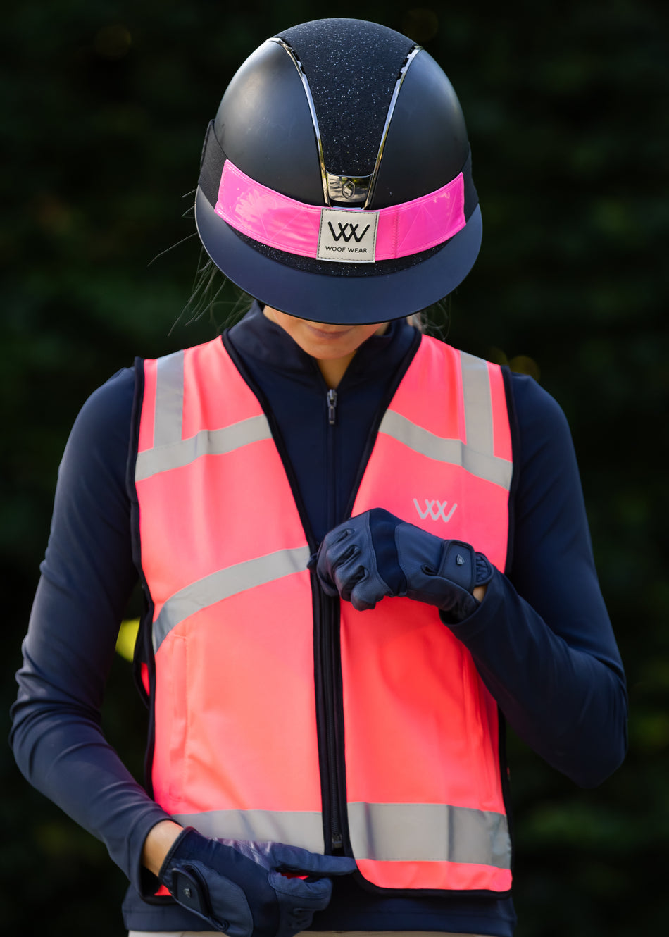 Person wearing a navy helmet with pink strap, reflective orange vest, and dark gloves against a blurred background.